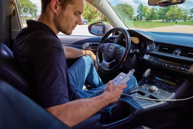 A man using an Intoxalock ignition interlock device in a car, preparing to take a breath sample